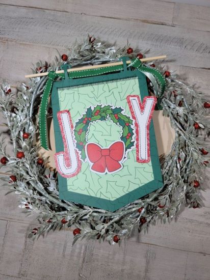 Die cut the base of the Big Banner 4 die in Spearmint and the smallest layer die in Mint Chip cardstock, glue them together. If you want do a zig zag stitch in the top part. Use the outline holly stamp and stamp at random to create a background.  Using the hollywreath2stamp dies, cut the holly wreath (I use an Exacto knife to cut the center of the wreath) and the bow and using Adhesive Foam glue it in the center of the banner. Die cut a letter J and a Y with the 4" alphabet and glue them flat.
Keywords: BannerDies